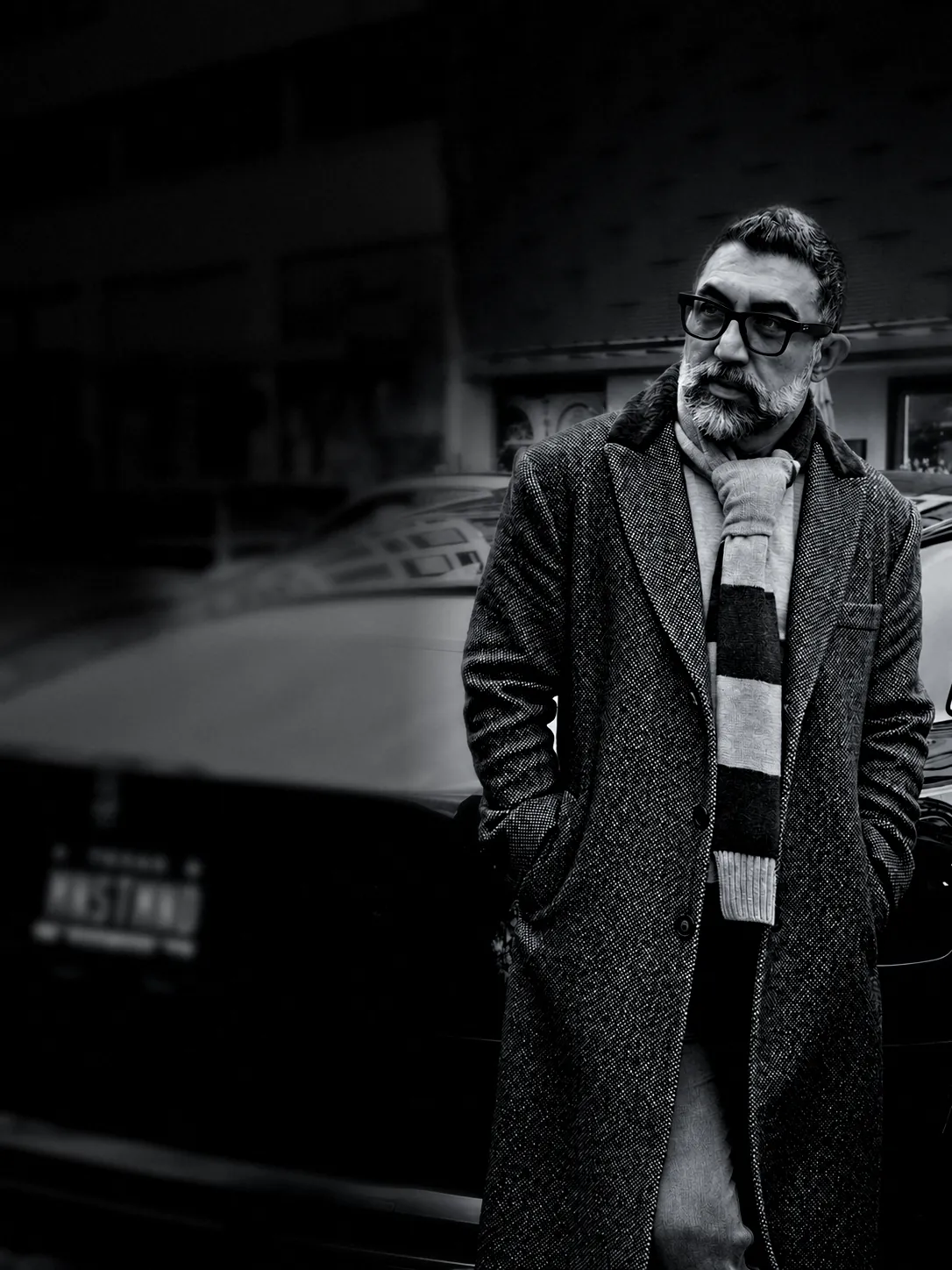 Houman Farzian in a grey overcoat and striped scarf beside a car with the MNSTMND plate, black and white
