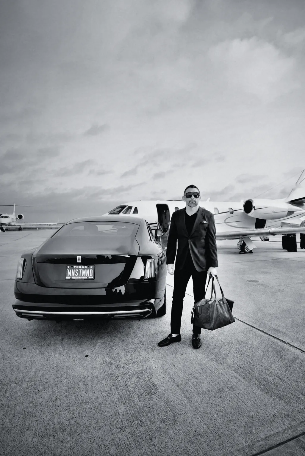 Houman Farzian on the tarmac between a car with the MNSTMND plate and a private jet, carrying a leather duffel, black and white